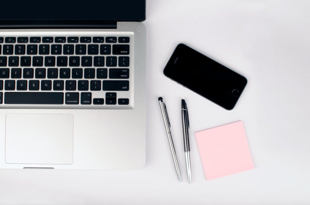 pexels photo 296115 A top view of a modern workspace featuring a laptop, smartphone, pens, and a pink sticky note on a white desk.