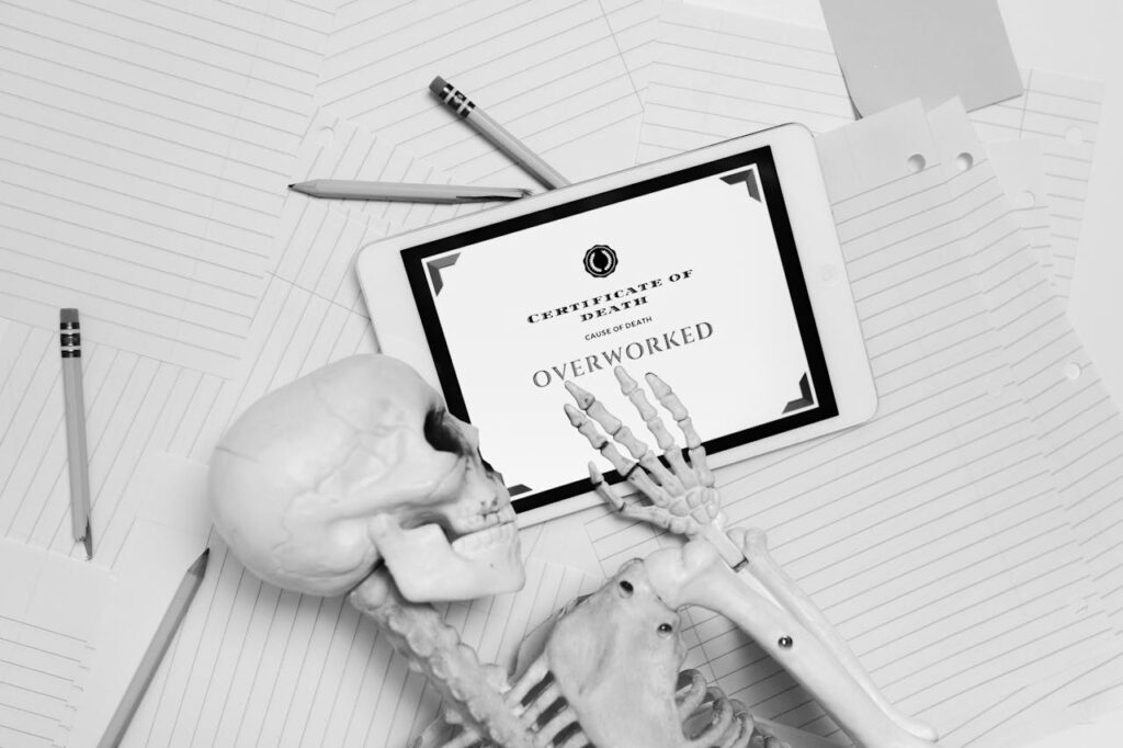pexels photo 8386713 Black and white image of skeleton hand holding tablet with 'Certificate of Death' showing overworked.